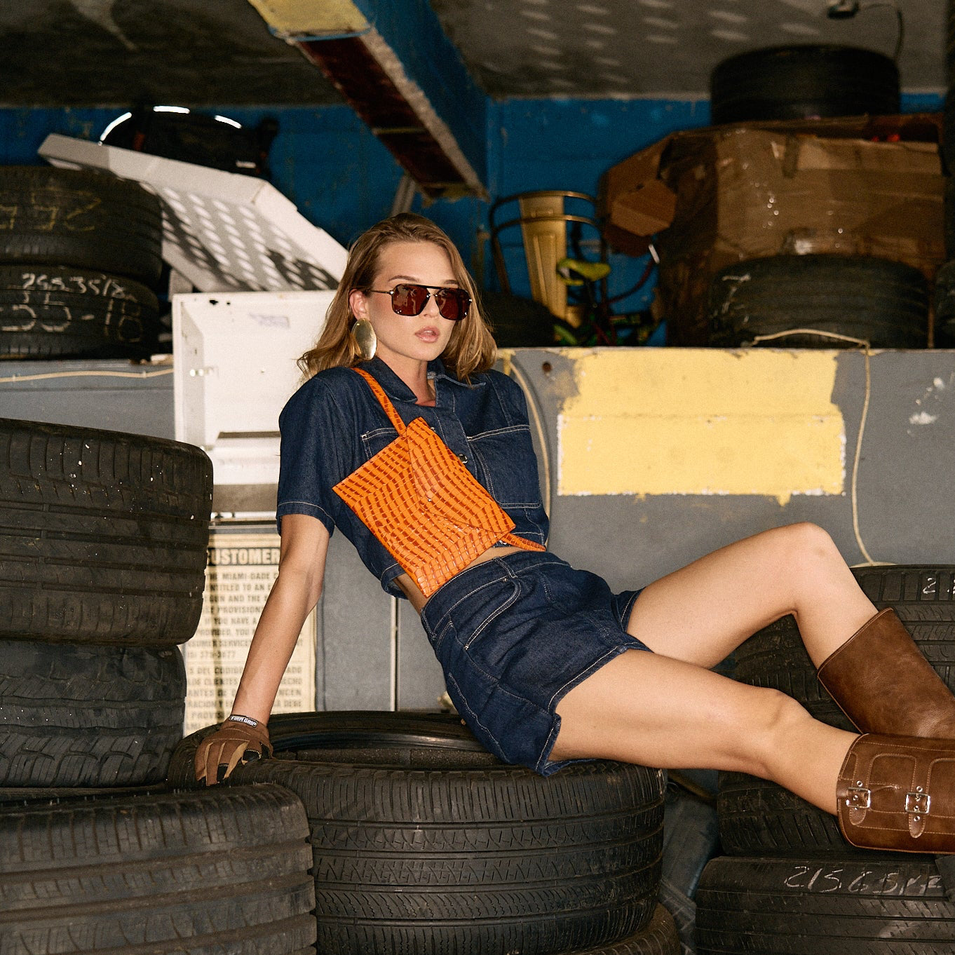 Woman wearing an orange crossbody bag sitting on tires in a warehouse setting