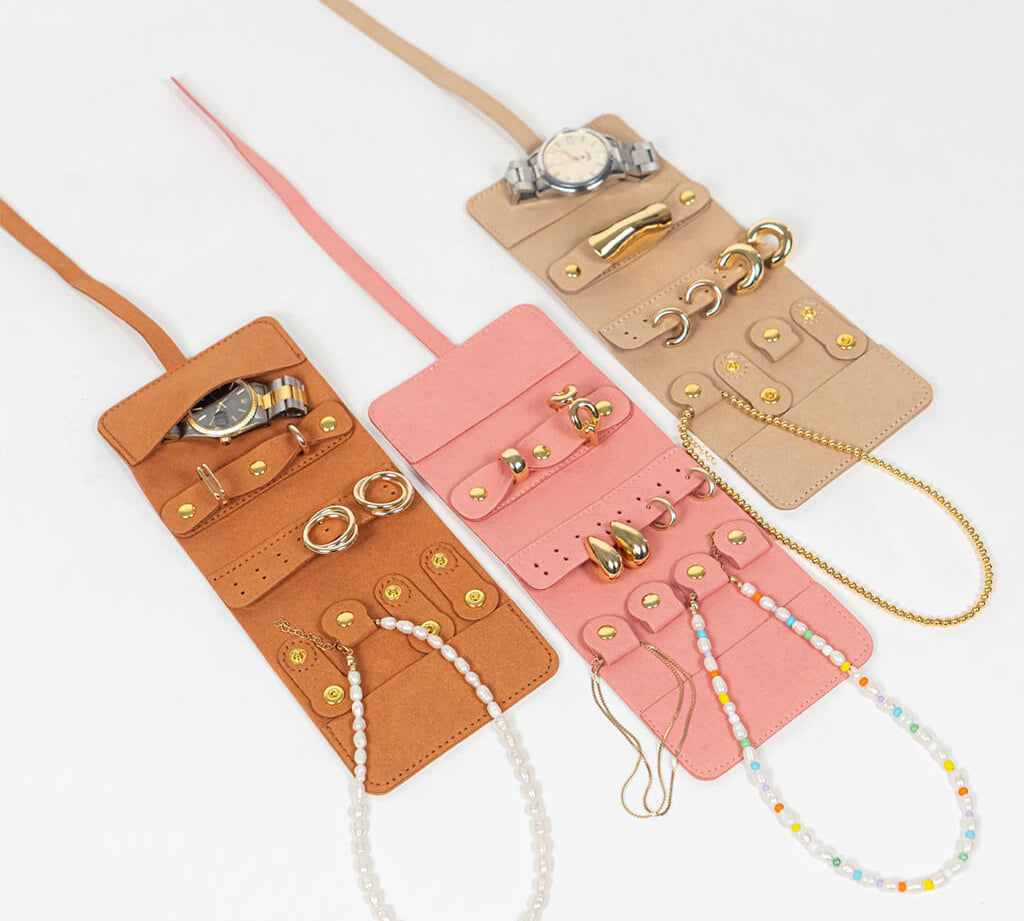 Three leather jewelry rolls in brown, pink, and beige with accessories on a white background